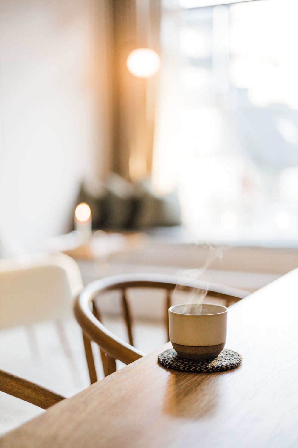 Steaming cup of coffee on wooden table with soft window light in background.
