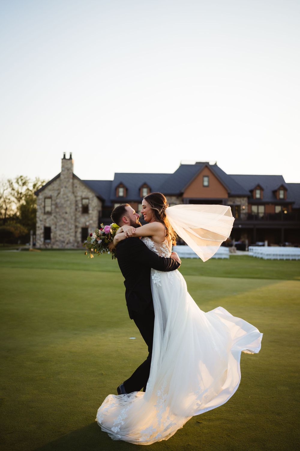 Couple embraces on a golf course with bride's dress flowing dramatically at sunset.