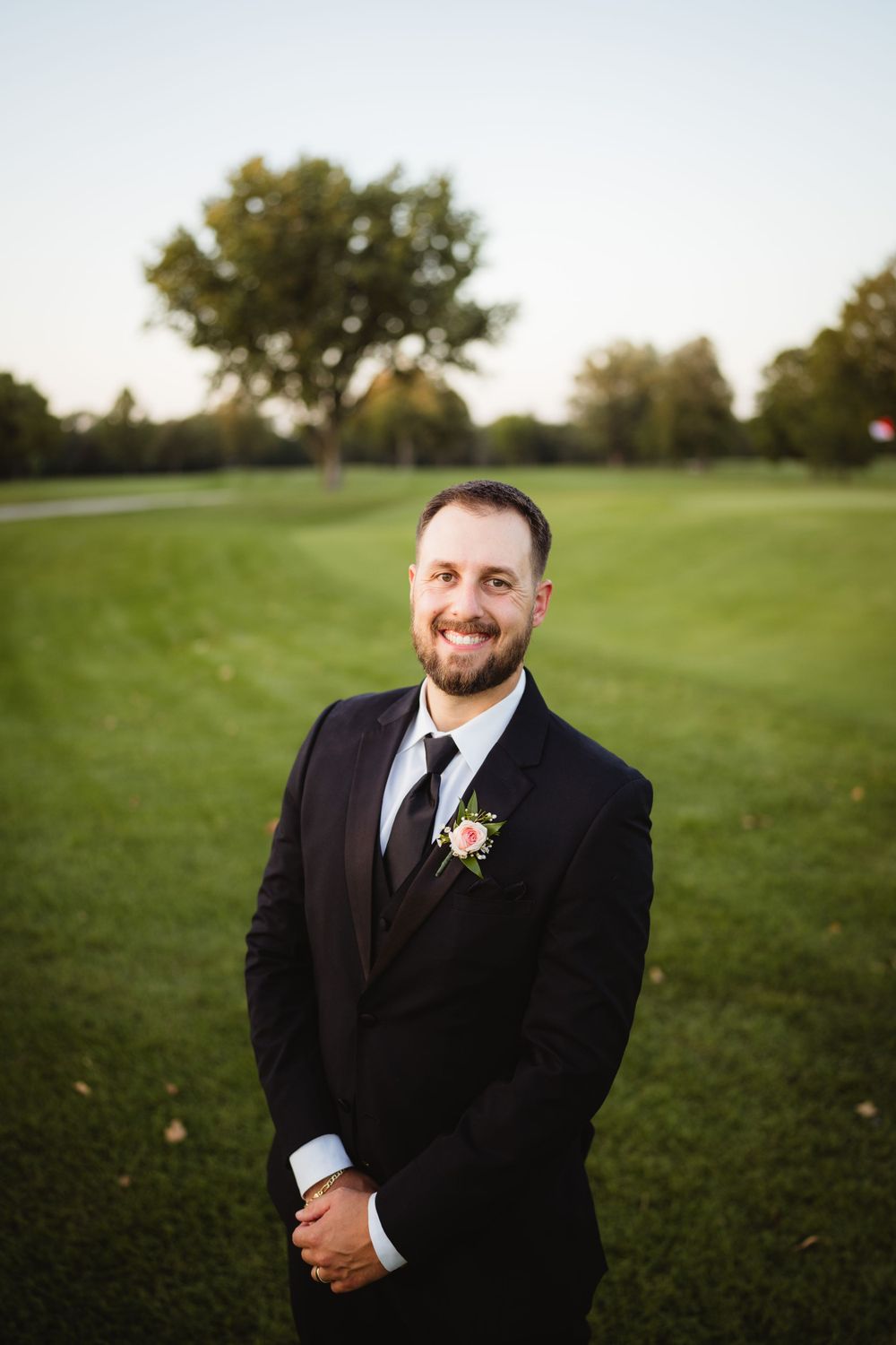 A person in a classic black tuxedo poses on a golf course at sunset.
