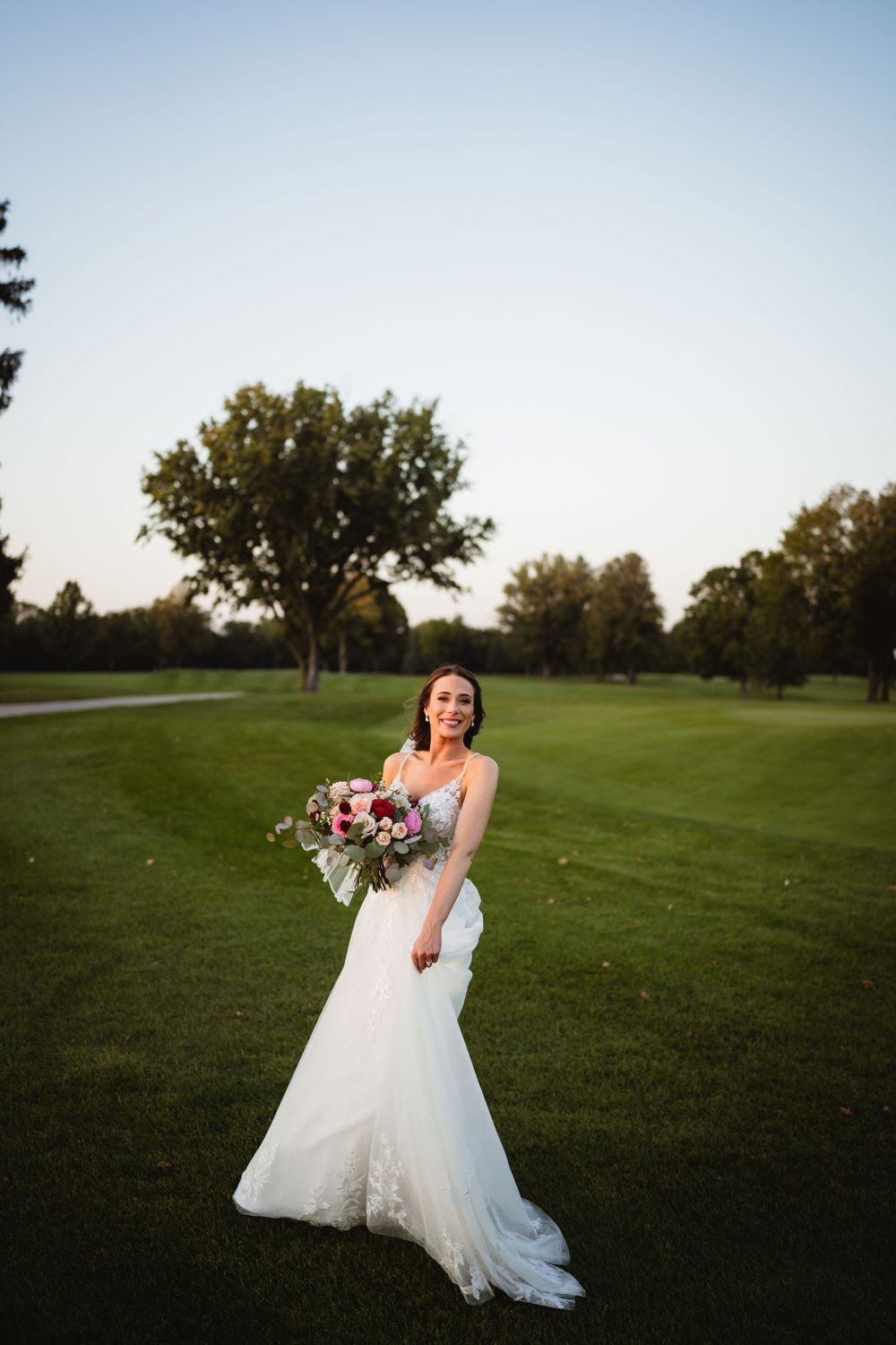 A bride in a flowing white gown poses with a bouquet on a golf course at sunset.