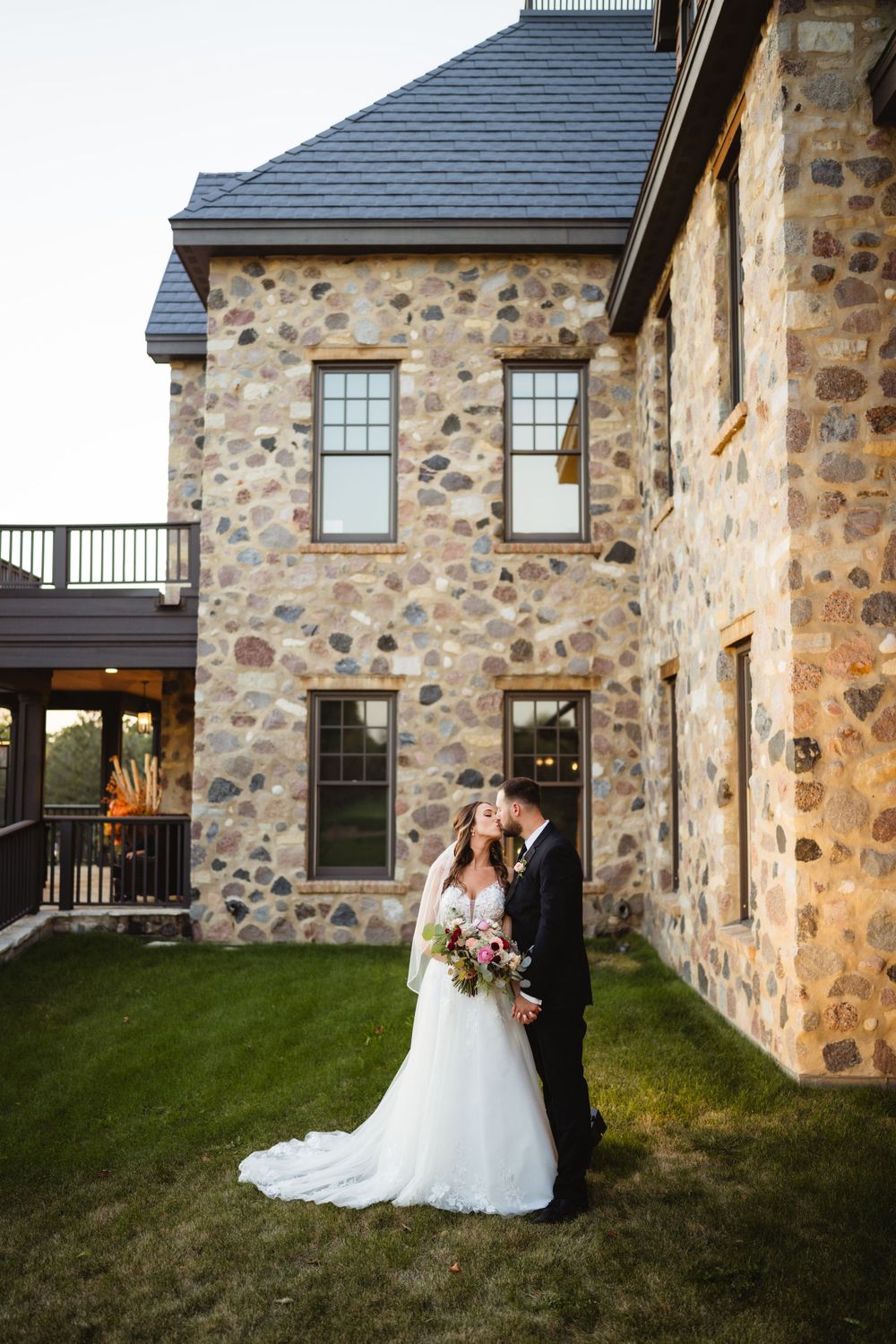 A couple poses beside a rustic stone building with slate roof during golden hour.