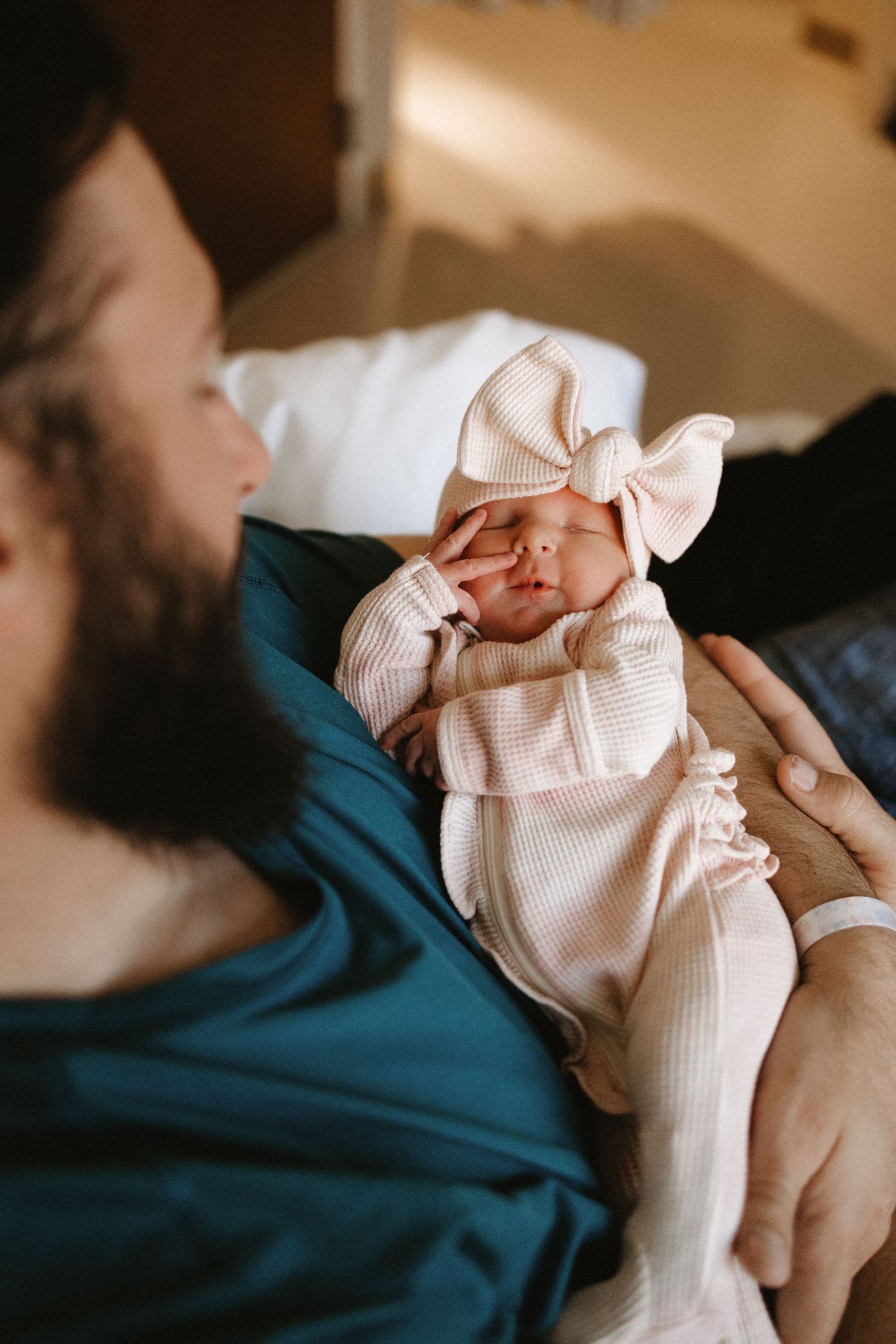 Baby wearing pink bow outfit being held by parent in blue shirt.