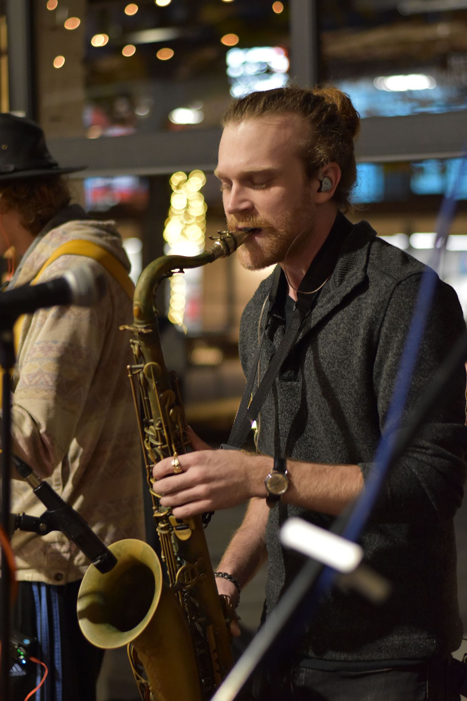 A jazz saxophonist performs on stage with a dark grey vest in a dimly lit venue with bokeh lights in the background.