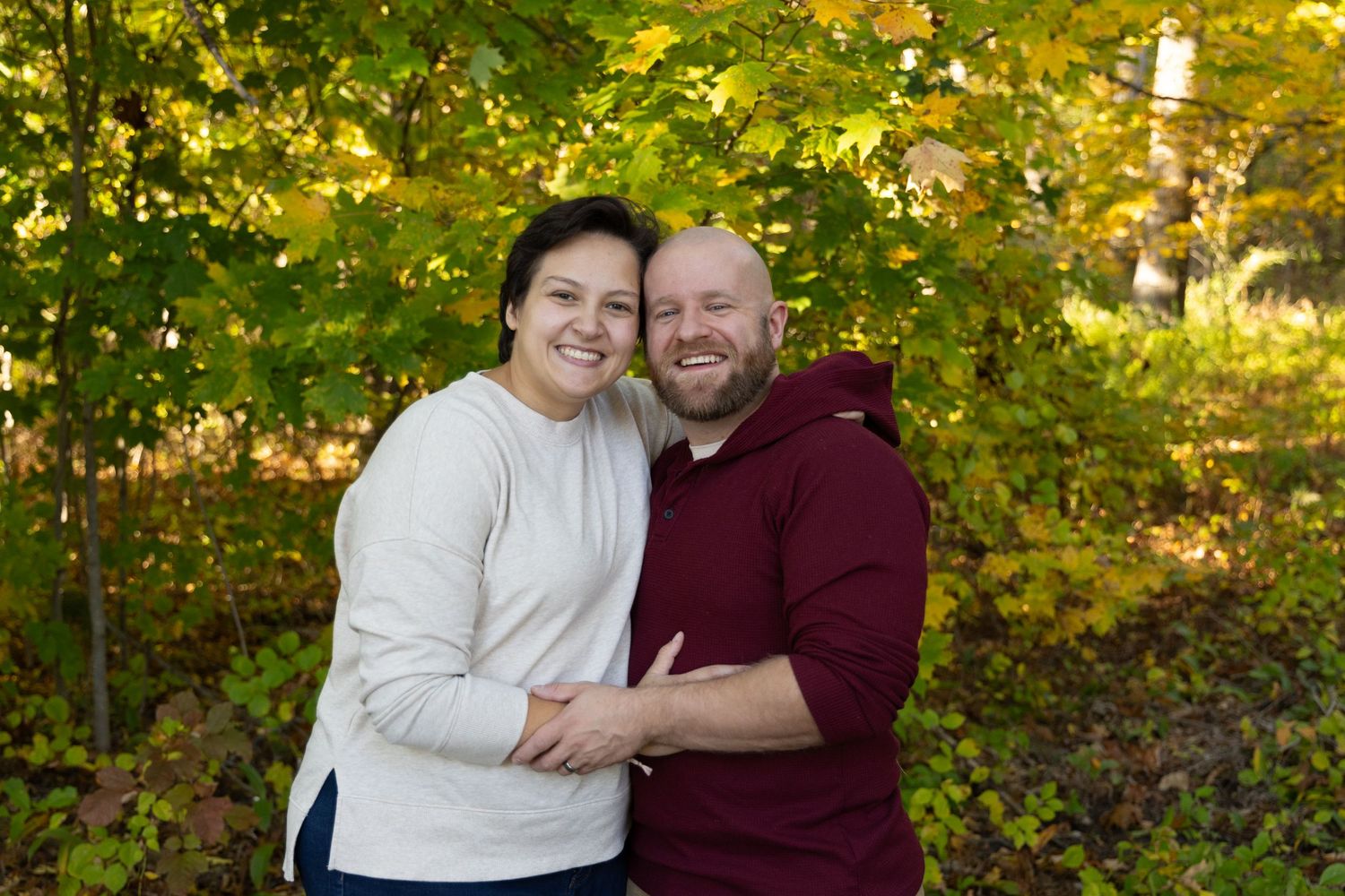Smiling couple embraces outdoors surrounded by vibrant autumn foliage in warm fall colors.