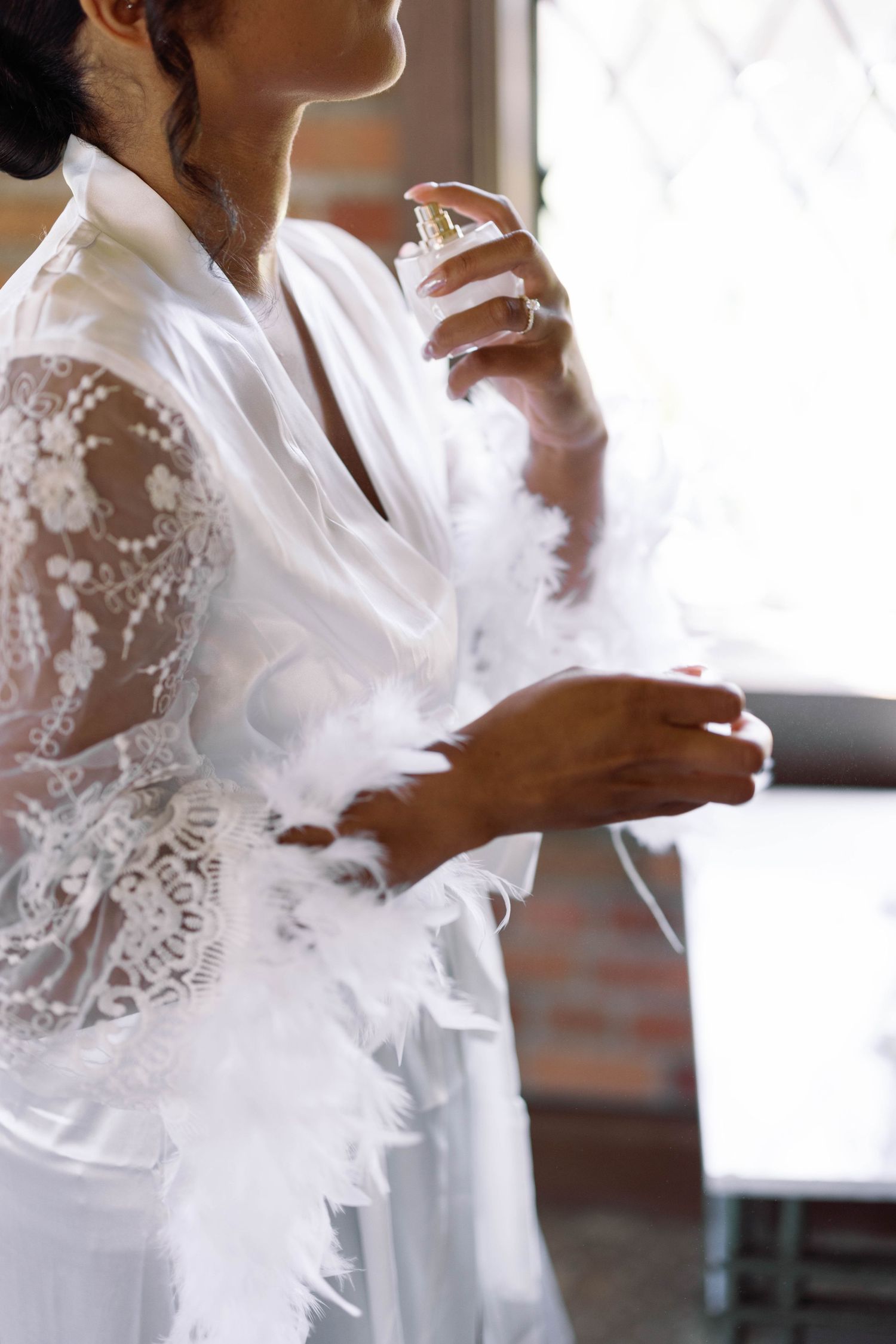 Person wearing delicate white lace robe sprays perfume in soft natural light.