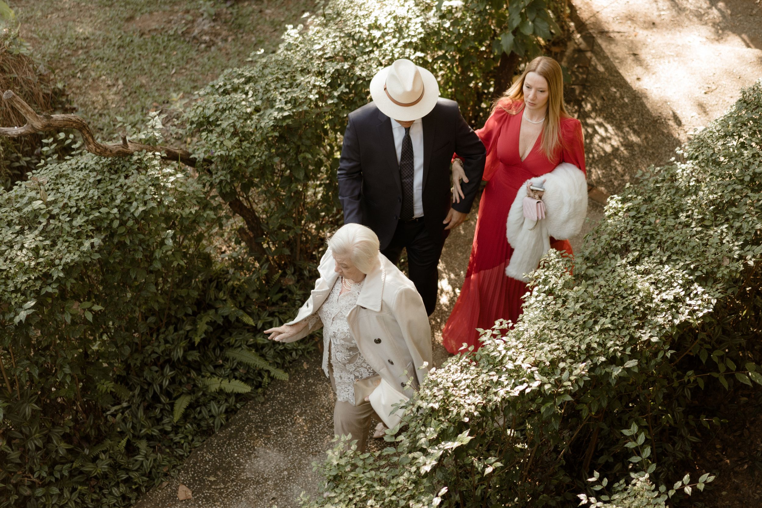 Garden party gathering with guests in white and red attire among blooming bushes.
