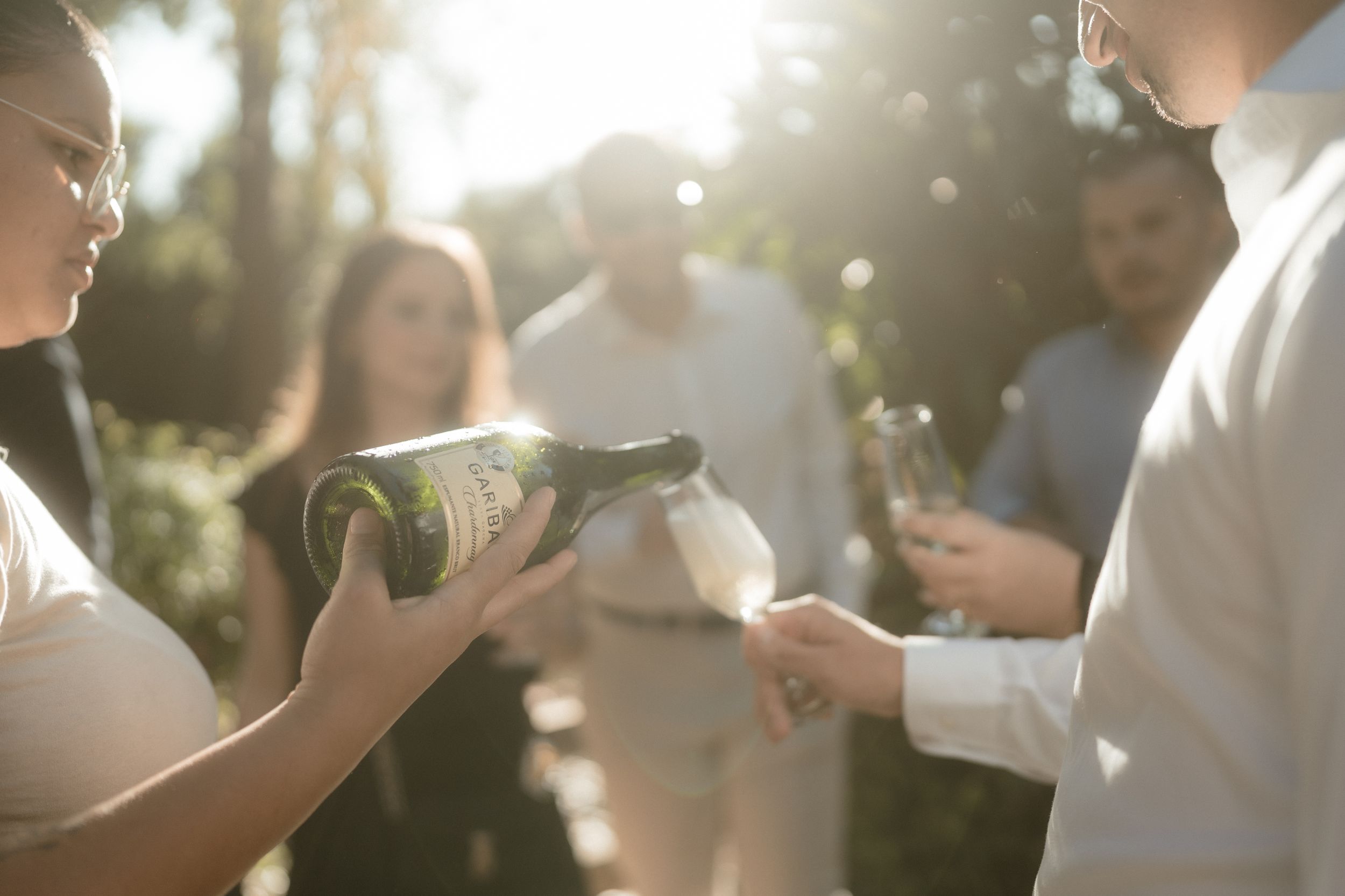Guests pouring champagne at an outdoor celebration with sunlight filtering through trees.