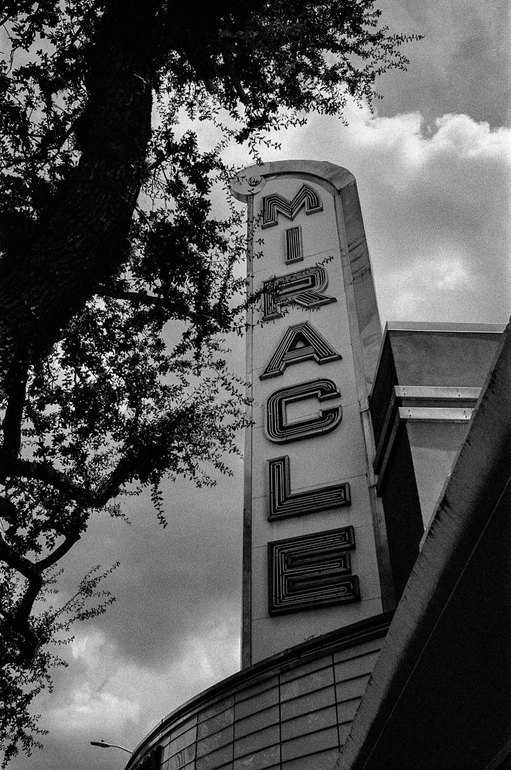 Vintage vertical Miracle theater sign photographed against cloudy sky.