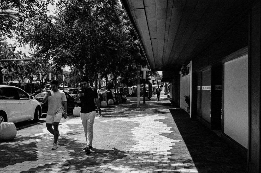 Pedestrians walking along covered sidewalk in front of urban storefronts.