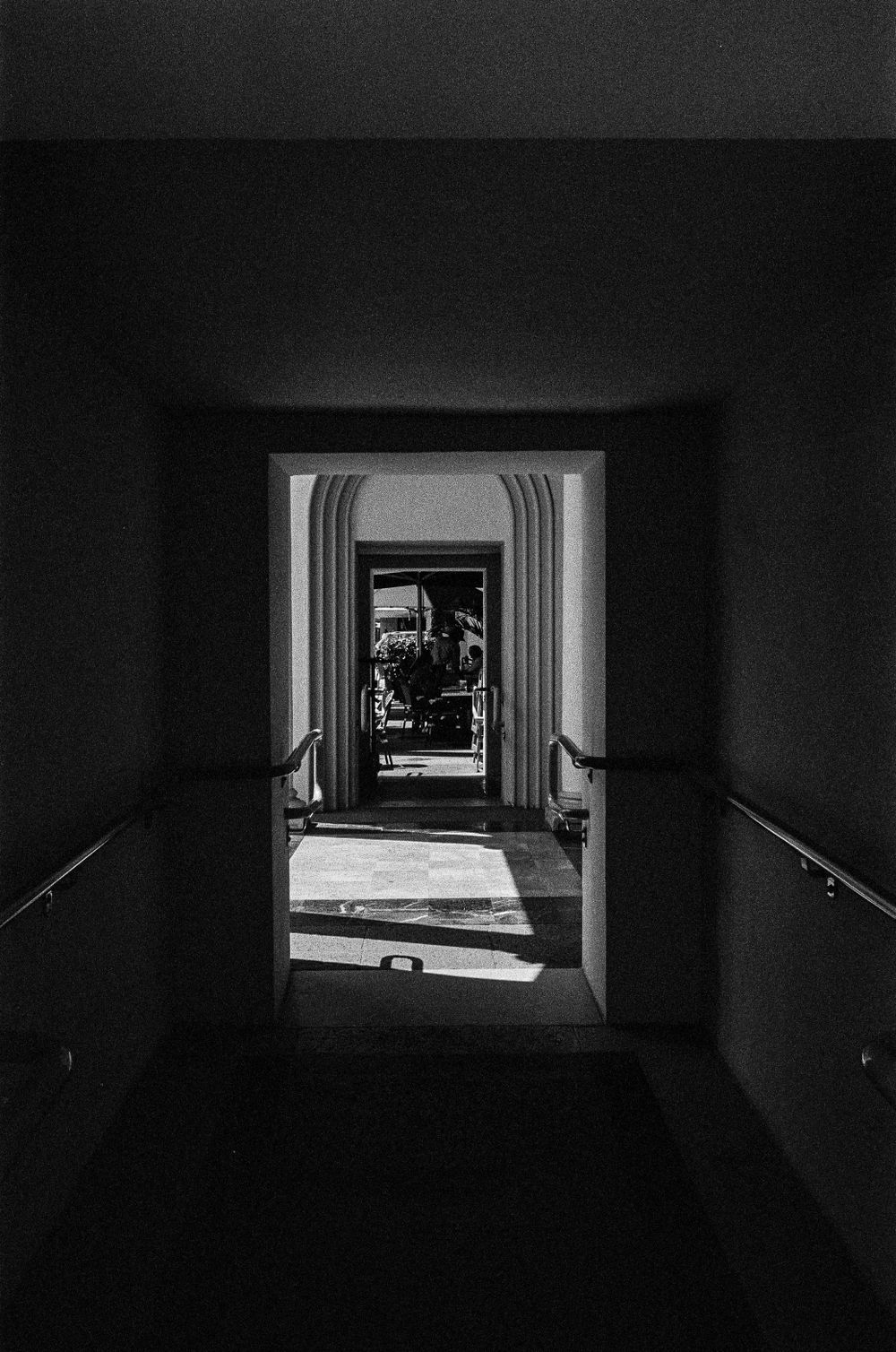 Dark hallway framed by handrails leads through multiple doorways creating a dramatic perspective in black and white.
