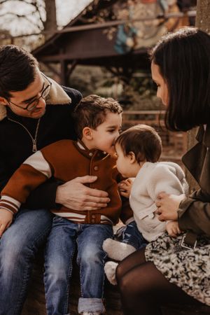Photo d'une famille lors d'une séance dans un parc. Un enfant embrasse son petit frère sur le front.