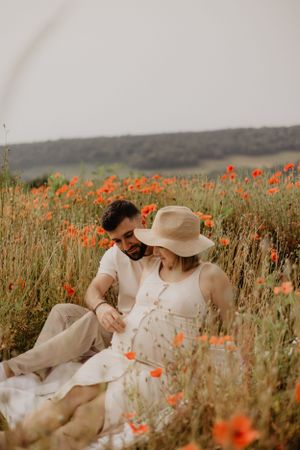 Photo d'un couple assis dans un champ de coquelicot. La femme est enceinte et l'homme touche son ventre.