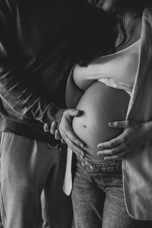 Photo en noir et blanc d'un ventre de femme enceinte. Son mari pose sa main sur le ventre de la femme enceinte.
