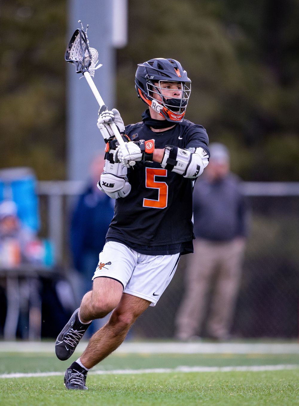 Lacrosse player in black jersey number 5 carries ball while running during competitive match.