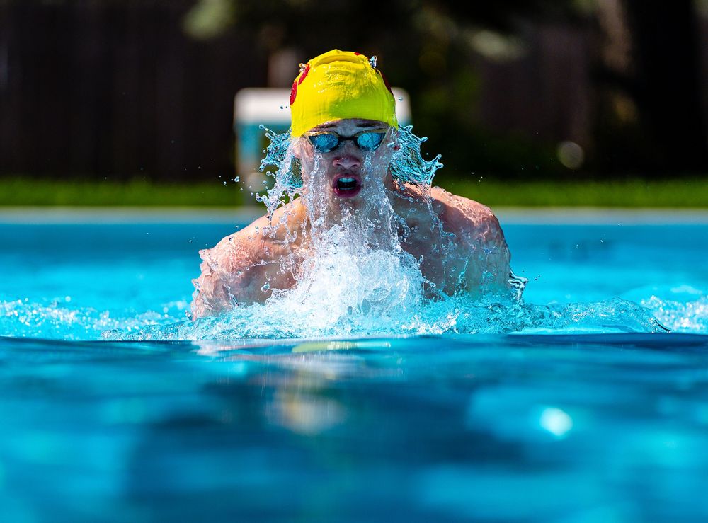 Competitive swimmer emerges from bright blue pool water while performing breaststroke wearing yellow swim cap.