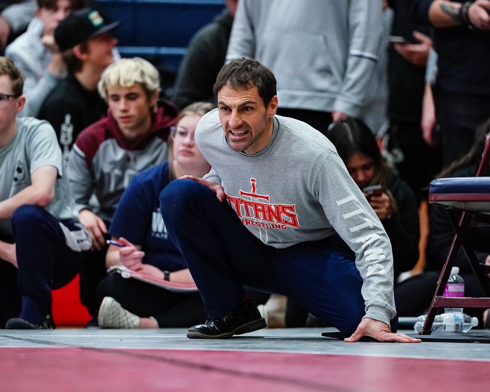 Wrestling coach in Titans sweatshirt observing match from crouched position.
