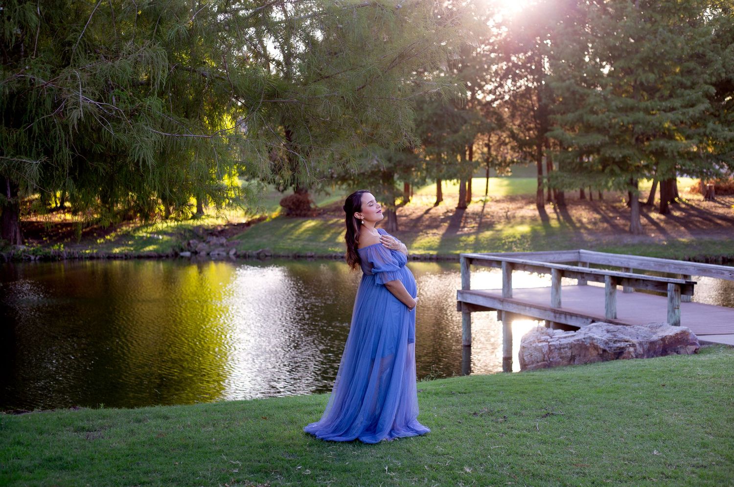 Pregnant woman in flowing blue gown poses gracefully by a lake at sunset near a wooden dock.