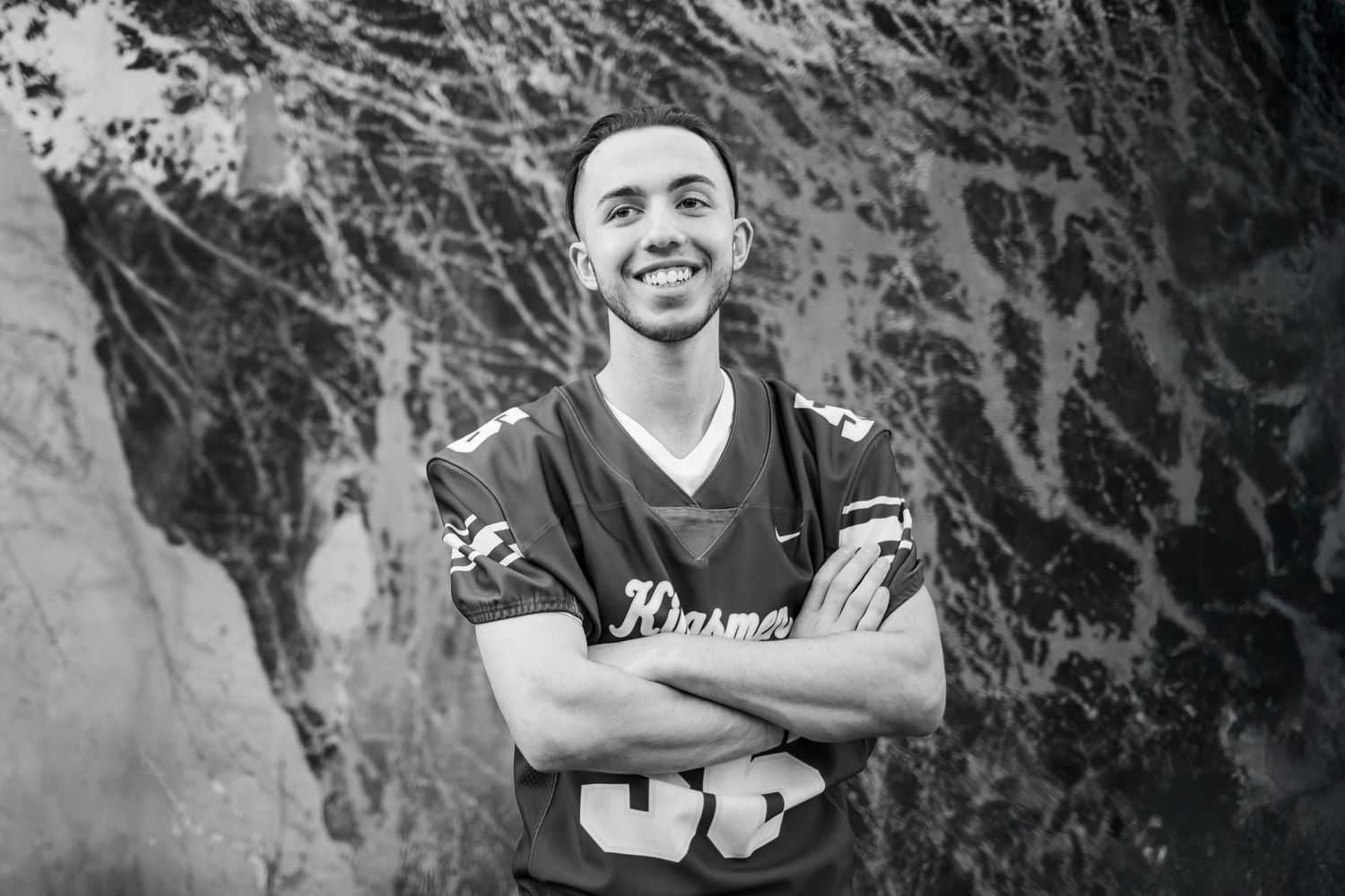 Black and white photo of someone in a dark sports jersey with arms crossed, smiling in front of a textured wall background.