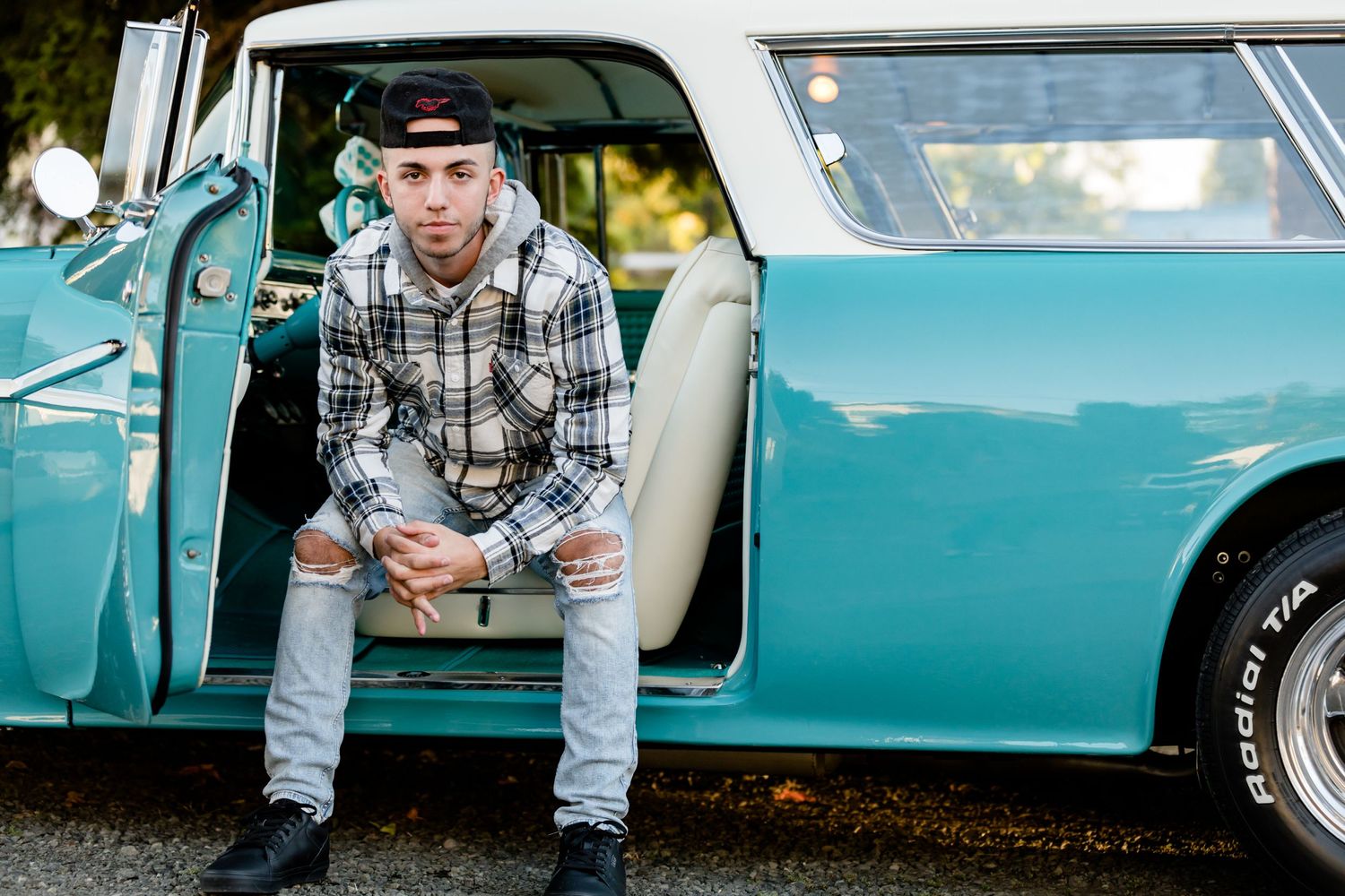 Person in plaid shirt and jeans sits in doorway of vintage turquoise classic car.
