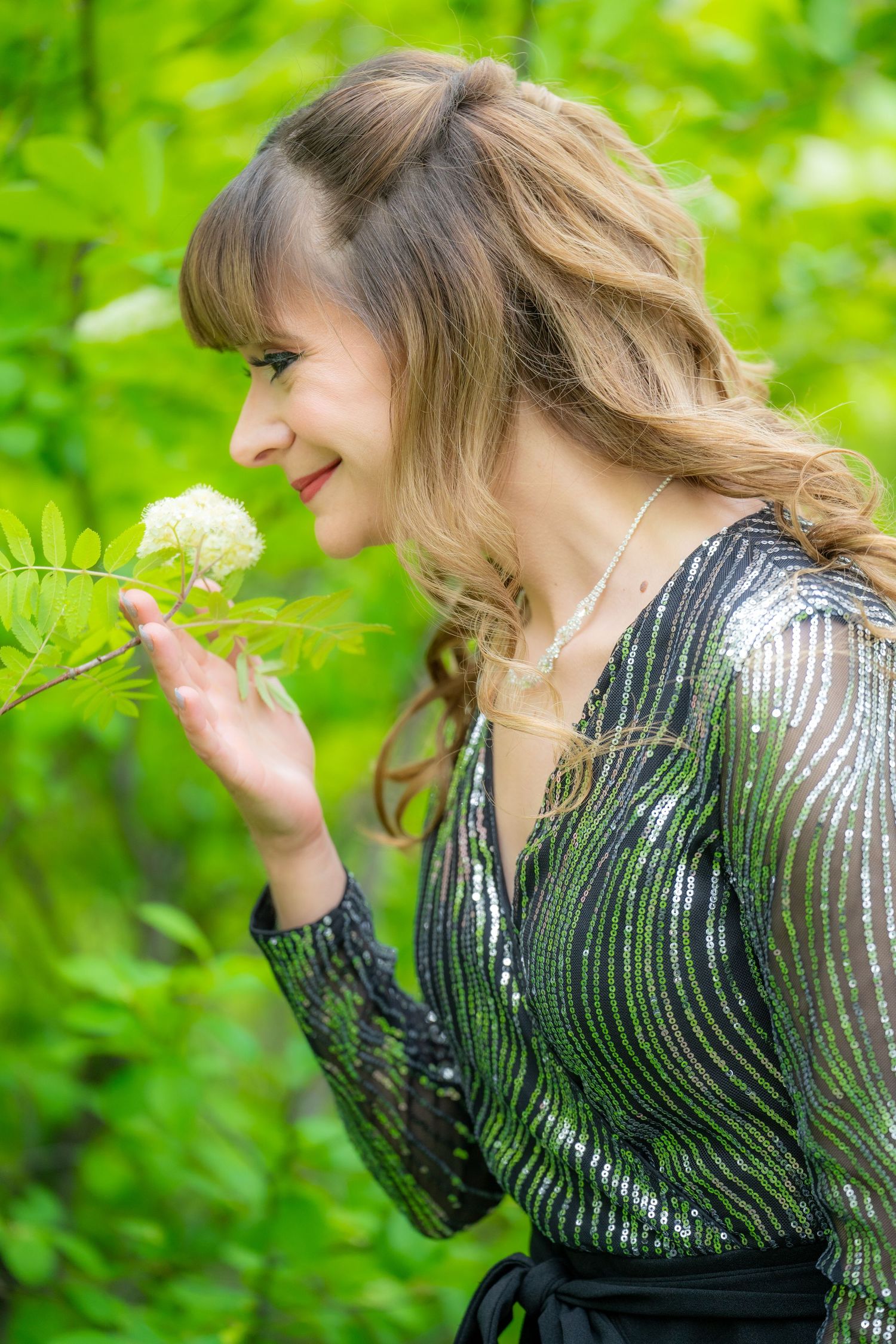 Side profile view of someone in a sparkly dress admiring a white dandelion flower against a green garden background.
