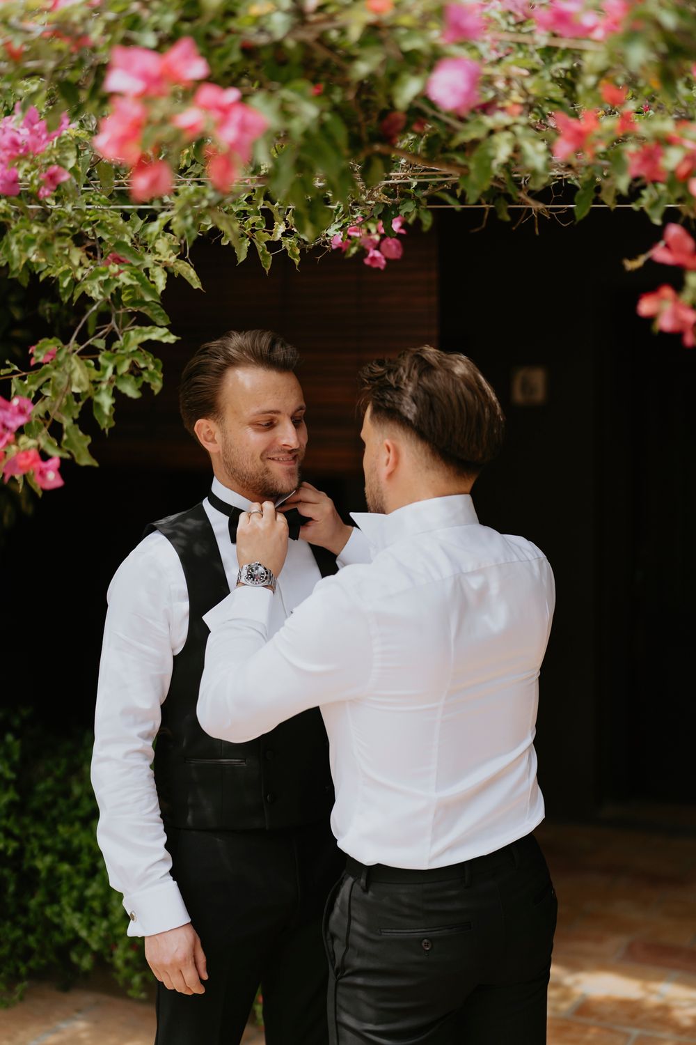 Romantic garden scene with pink bougainvillea flowers framing two people in formal attire adjusting a bow tie.