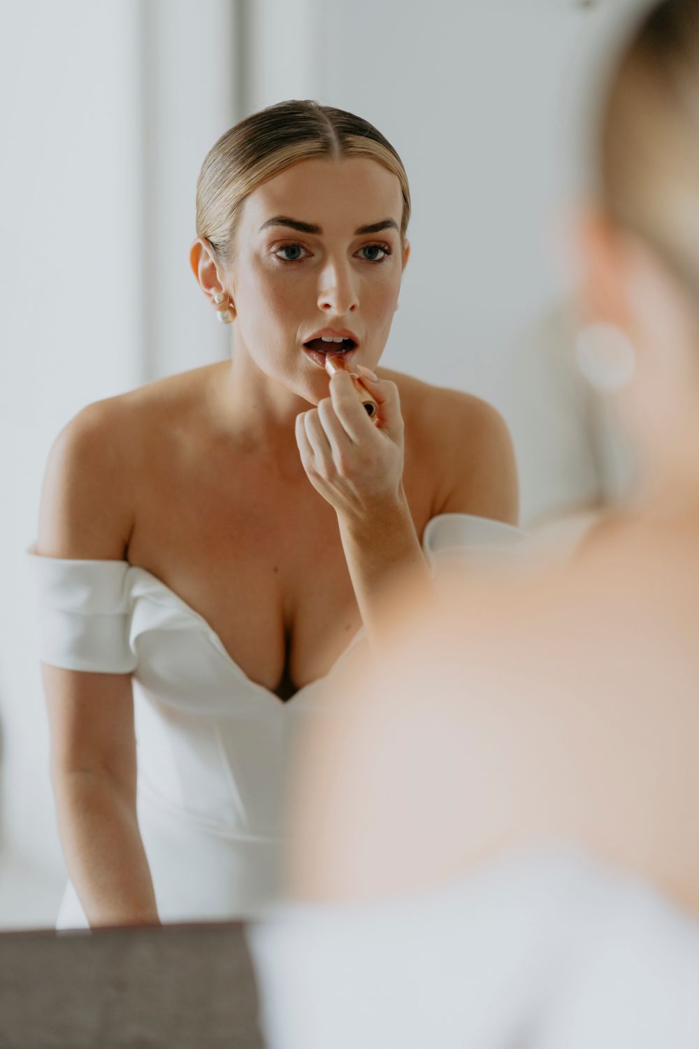 Mirror reflection of someone in a white off-shoulder dress applying lipstick while getting ready.