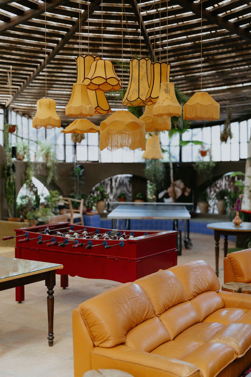 Modern game room with yellow pendant lamps, red foosball table and tan leather couch under a slatted roof.