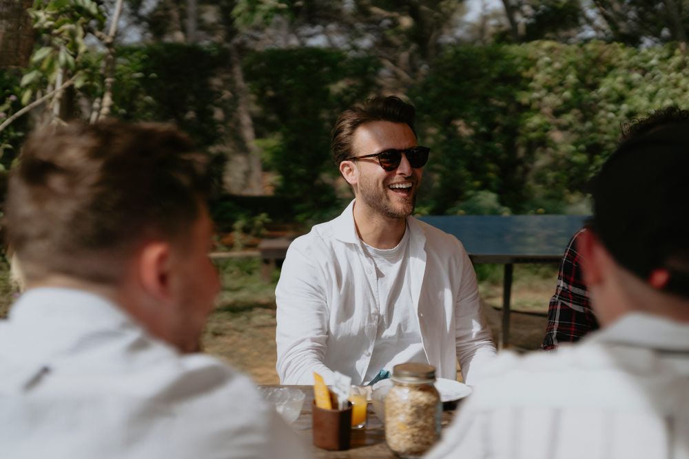 Three people sit at an outdoor table having drinks and conversation on a sunny day.