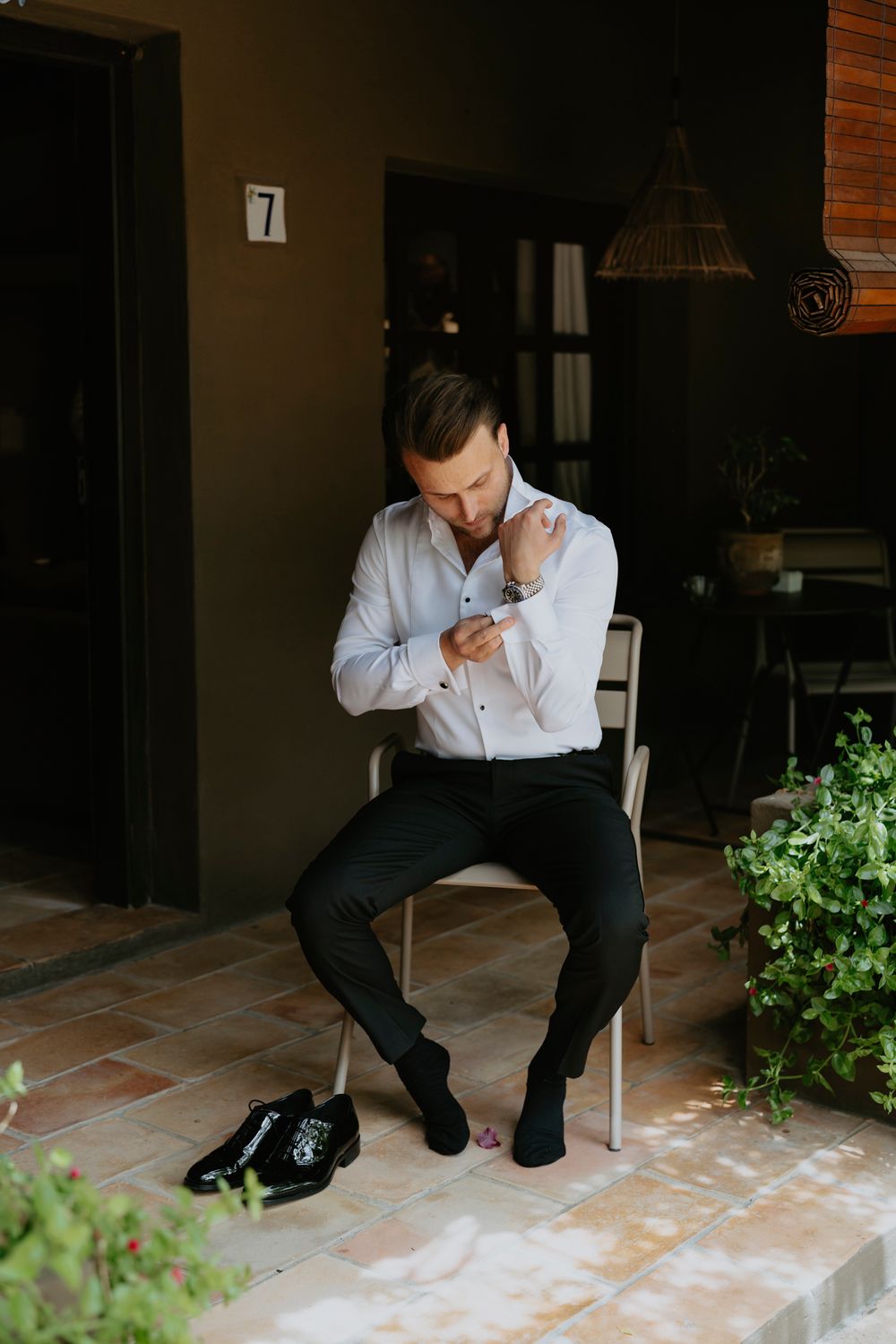 Someone straightening their light blue dress shirt while sitting on a concrete porch with black dress shoes nearby.