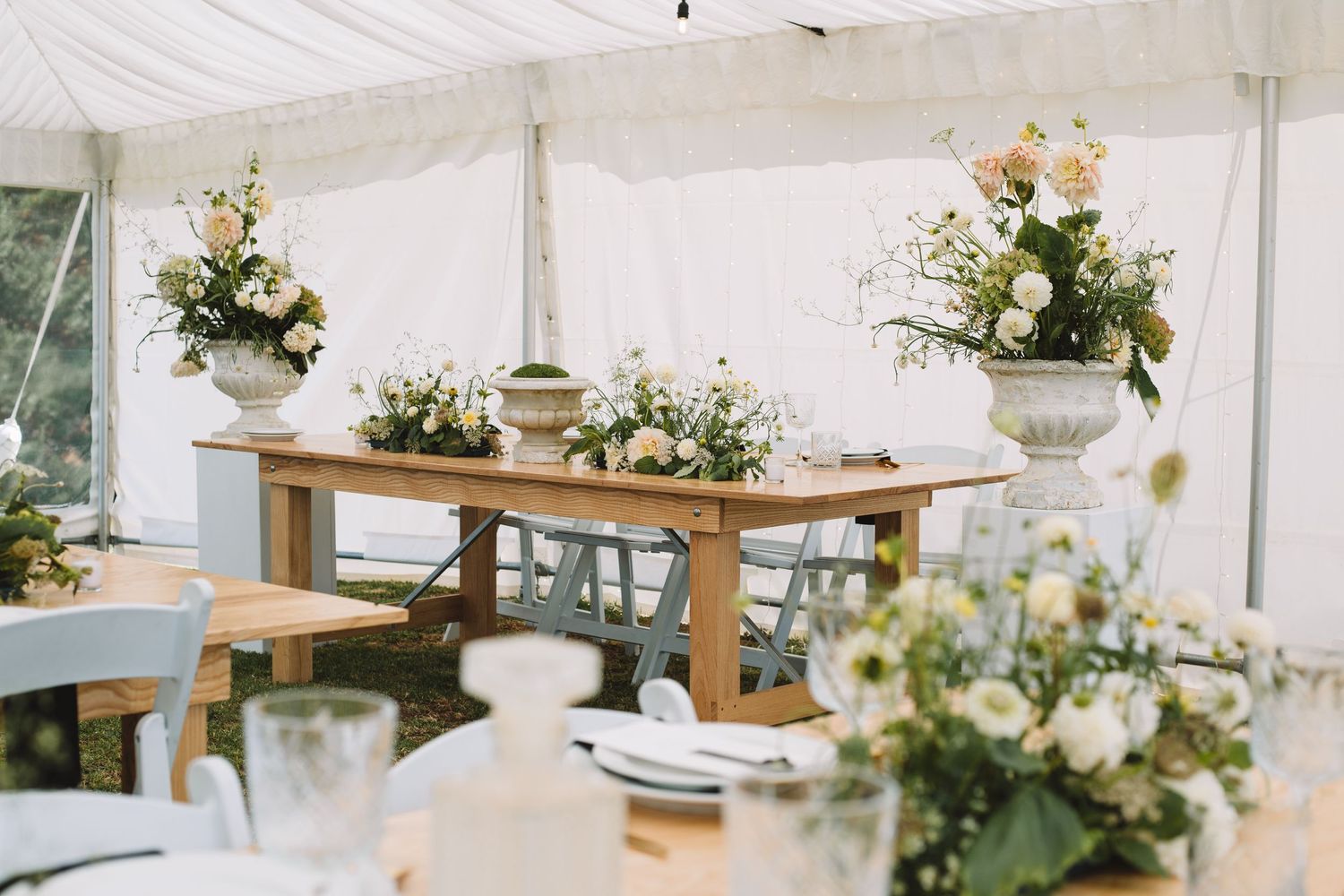 Elegant wedding reception tent with rustic wooden tables and floral centerpieces in white and green arrangements.