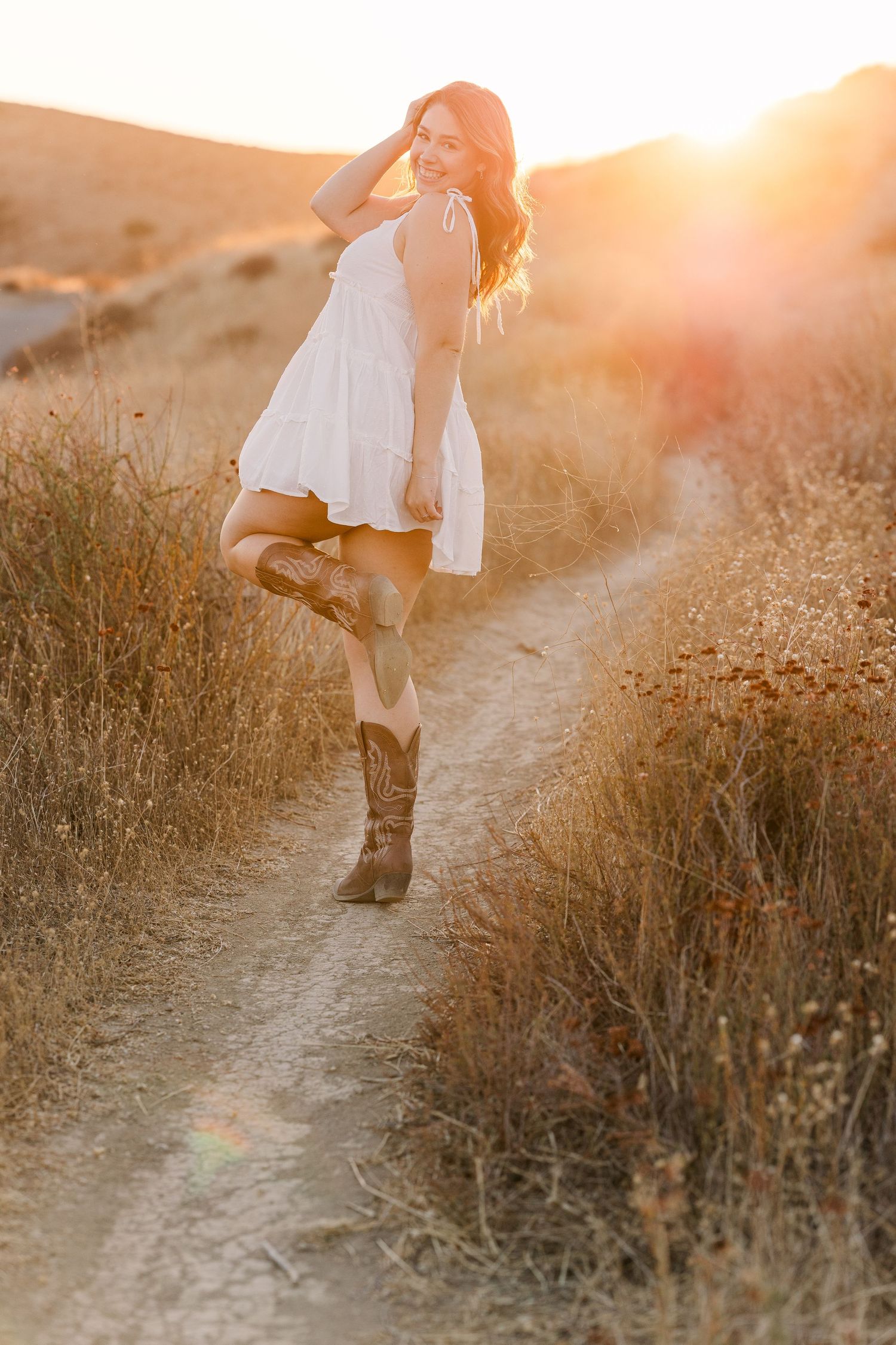 A white summer dress and boots outfit captured on a dirt trail during golden hour sunset with lens flare.