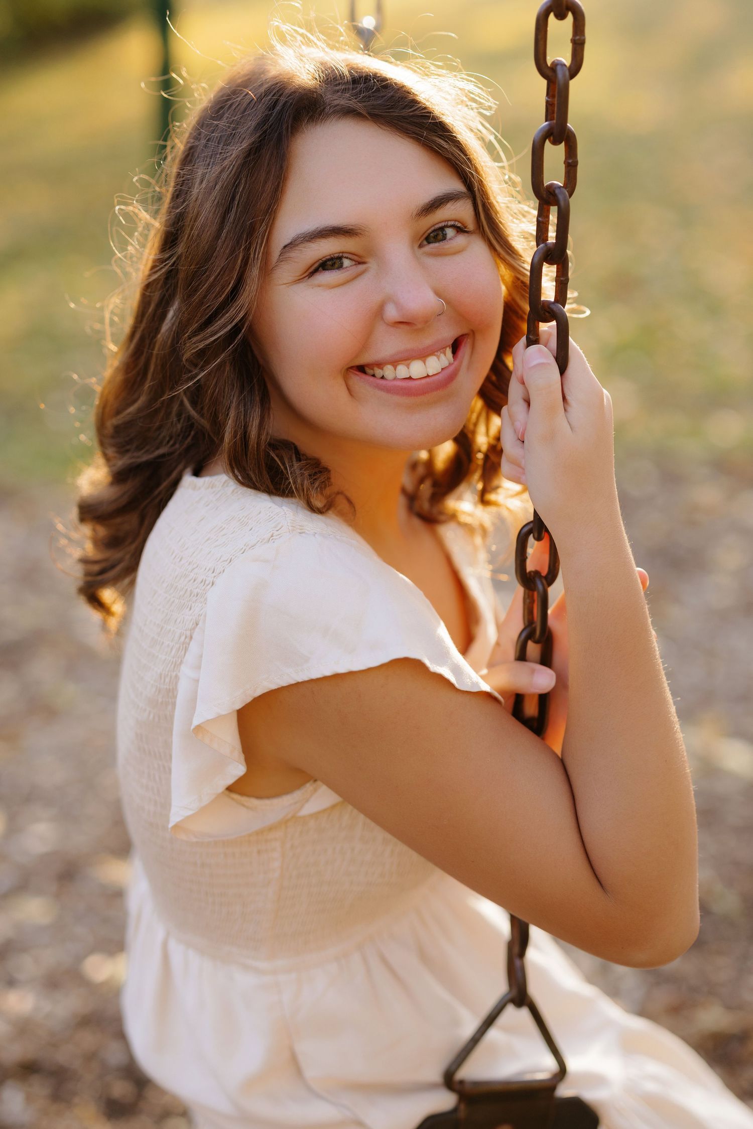 A joyful portrait of someone in a white summer dress holding a rustic chain swing in warm golden sunlight.