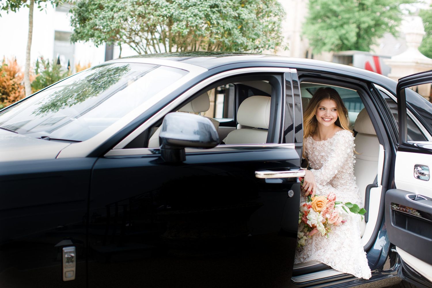 A bride wearing a white lace dress sits in a black luxury wedding car holding a pink and white flower bouquet in Halifax, Nova Scotia