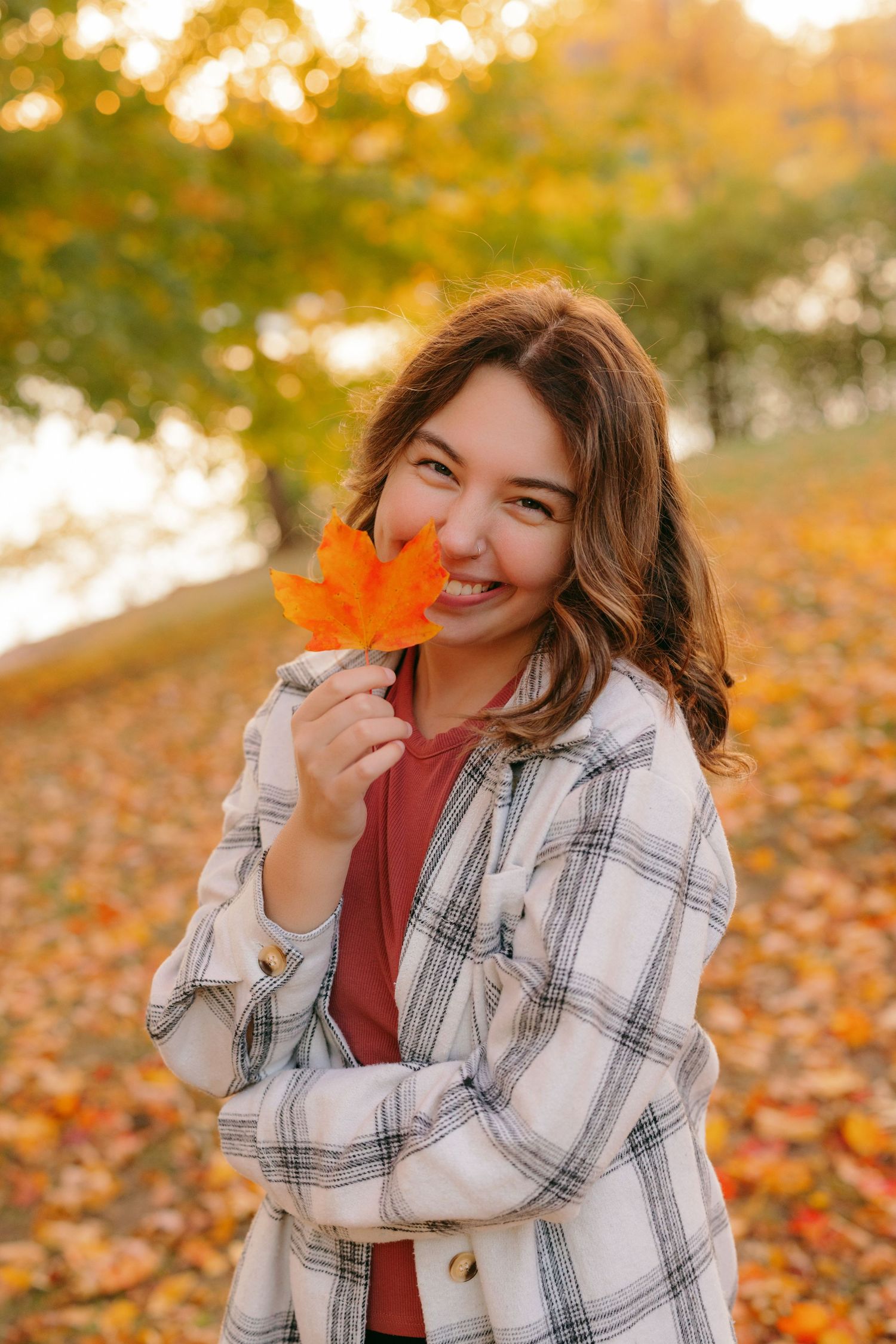 A stylish plaid jacket outfit for fall with an orange maple leaf against a backdrop of autumn foliage and golden light.