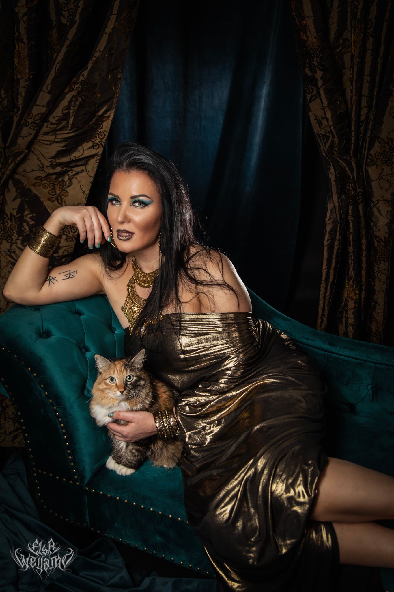 A glamorous photoshoot on a teal velvet sofa features a gold evening dress and orange cat in dark moody lighting.