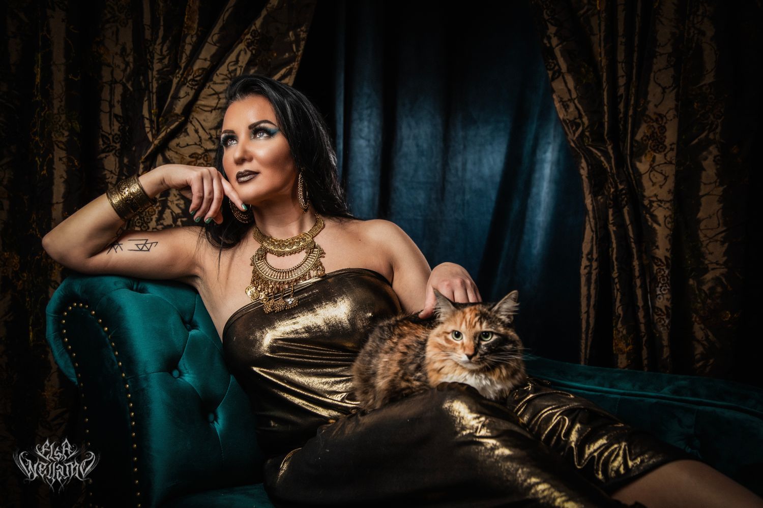 Glamorous portrait of woman in gold dress reclining on teal velvet chaise lounge with cat companion in moody lighting.