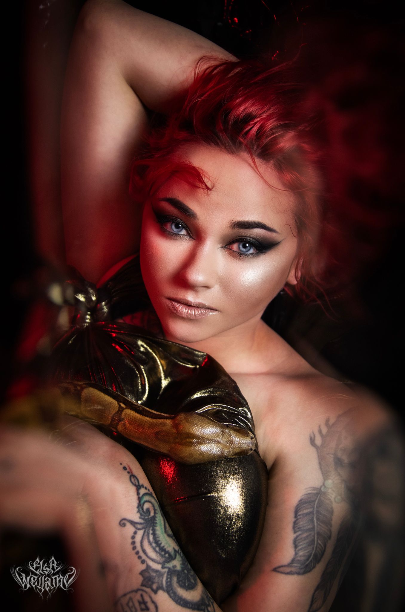 Artistic fashion portrait featuring vibrant red hair, dramatic makeup, gold gloves and visible tattoos in moody lighting.