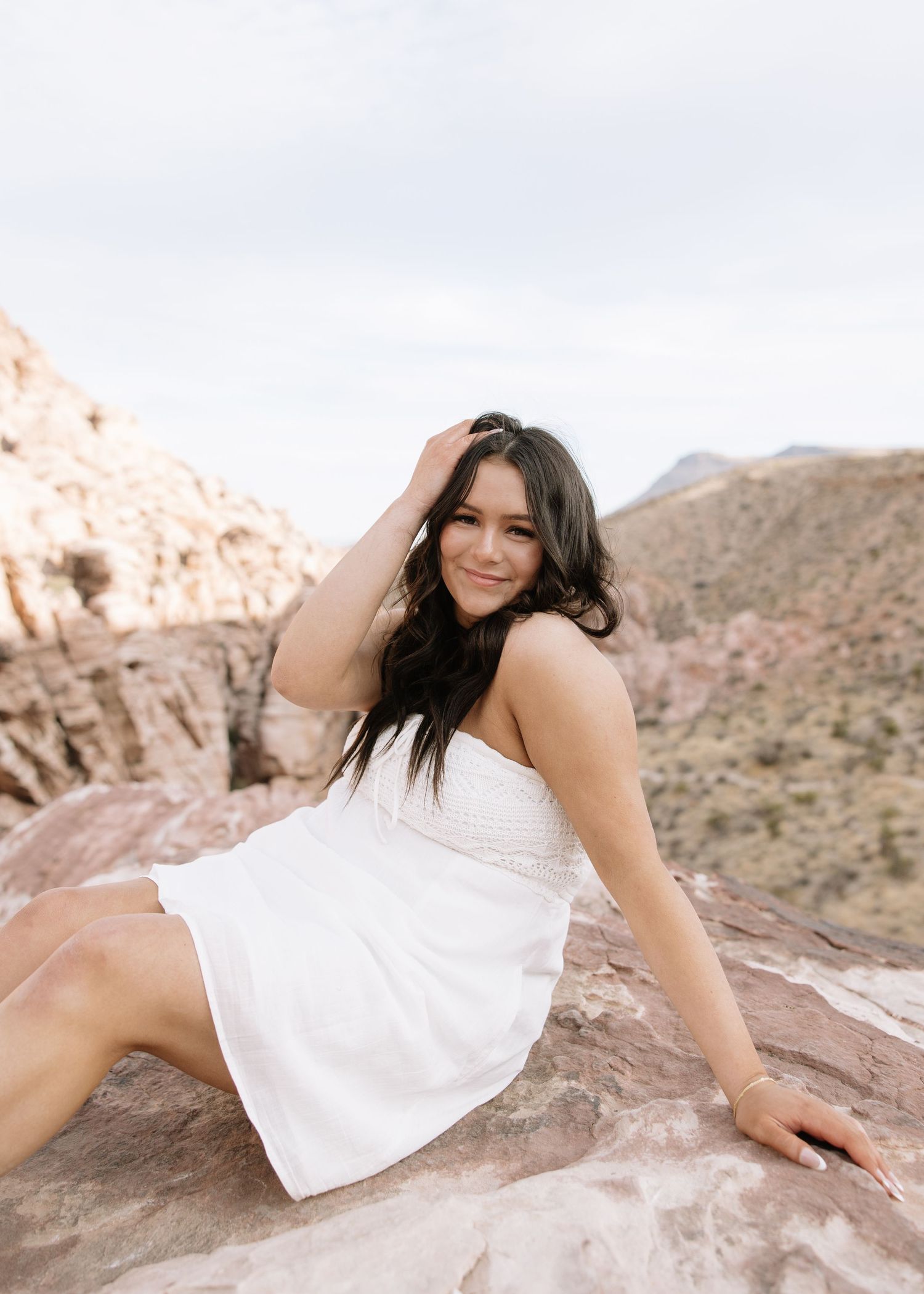 Person in white dress poses gracefully on red rock formations in desert mountain landscape.