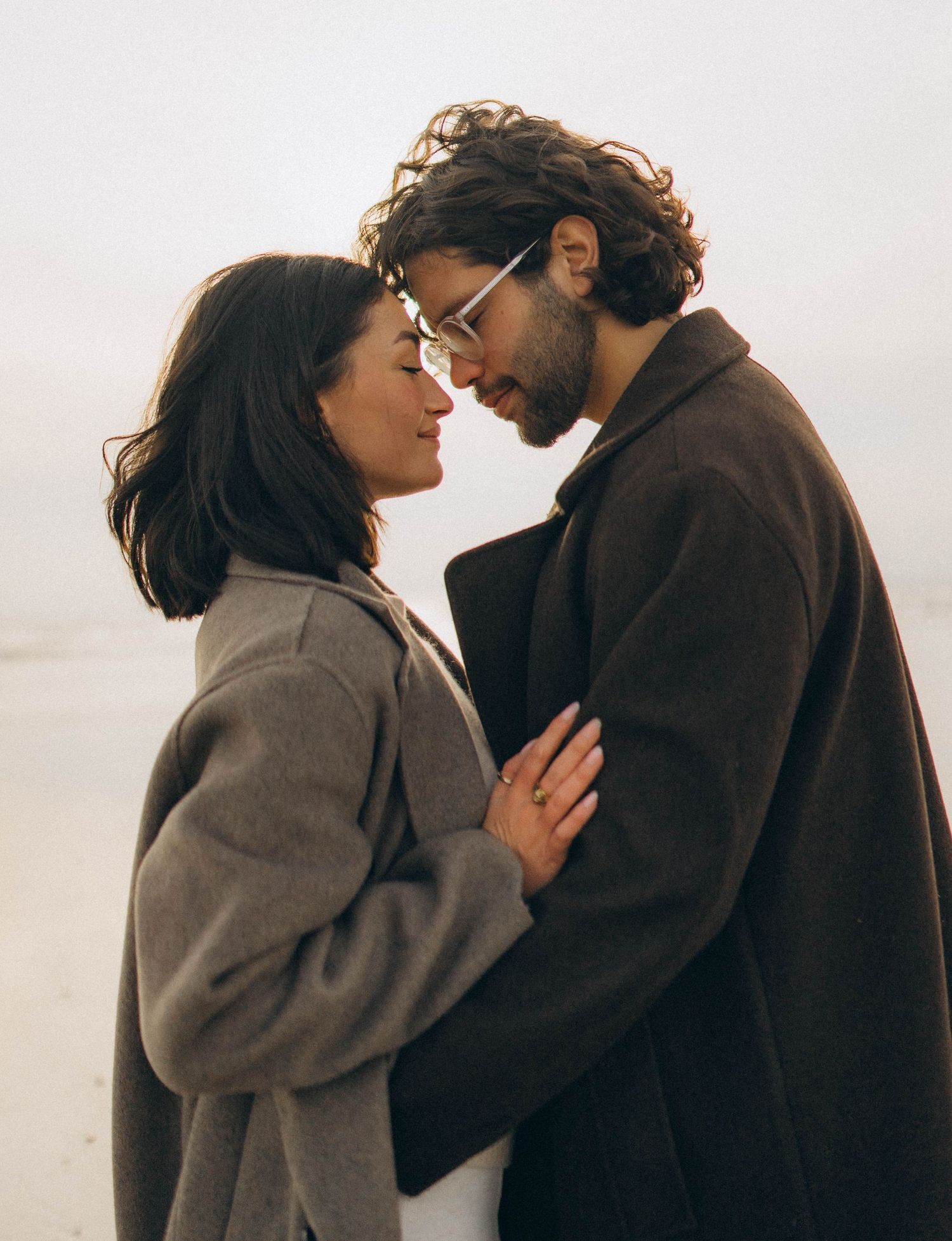 A couple sharing intimate moments against a light background wearing cozy winter coats.