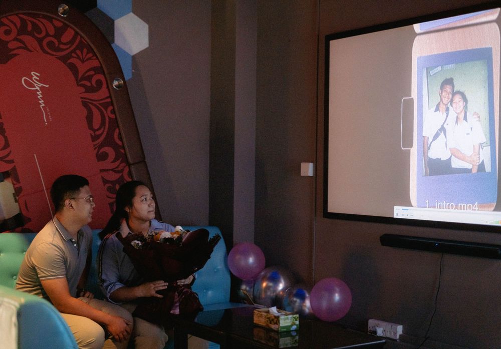 People watching a presentation on a large screen in a dimly lit room with purple balloons.