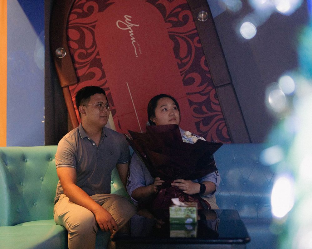 A couple sits on a plush couch in a colorfully lit room with decorative wall features.
