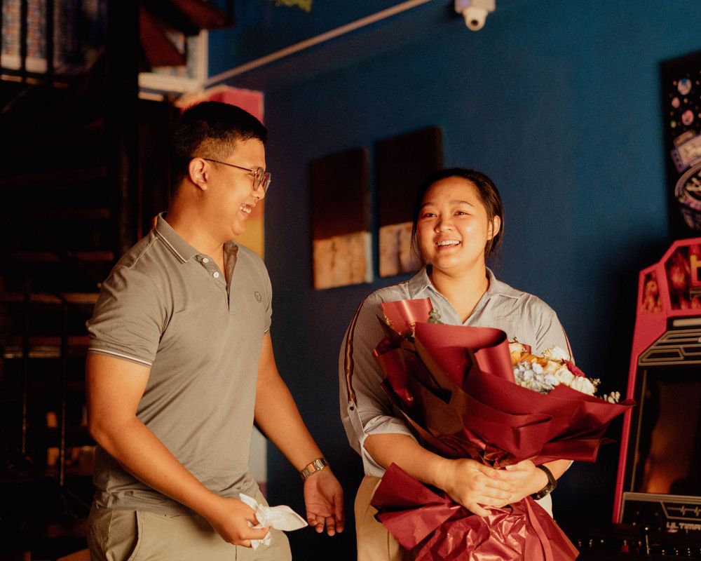 A couple shares a joyful moment while holding a red bouquet in a blue-walled room.