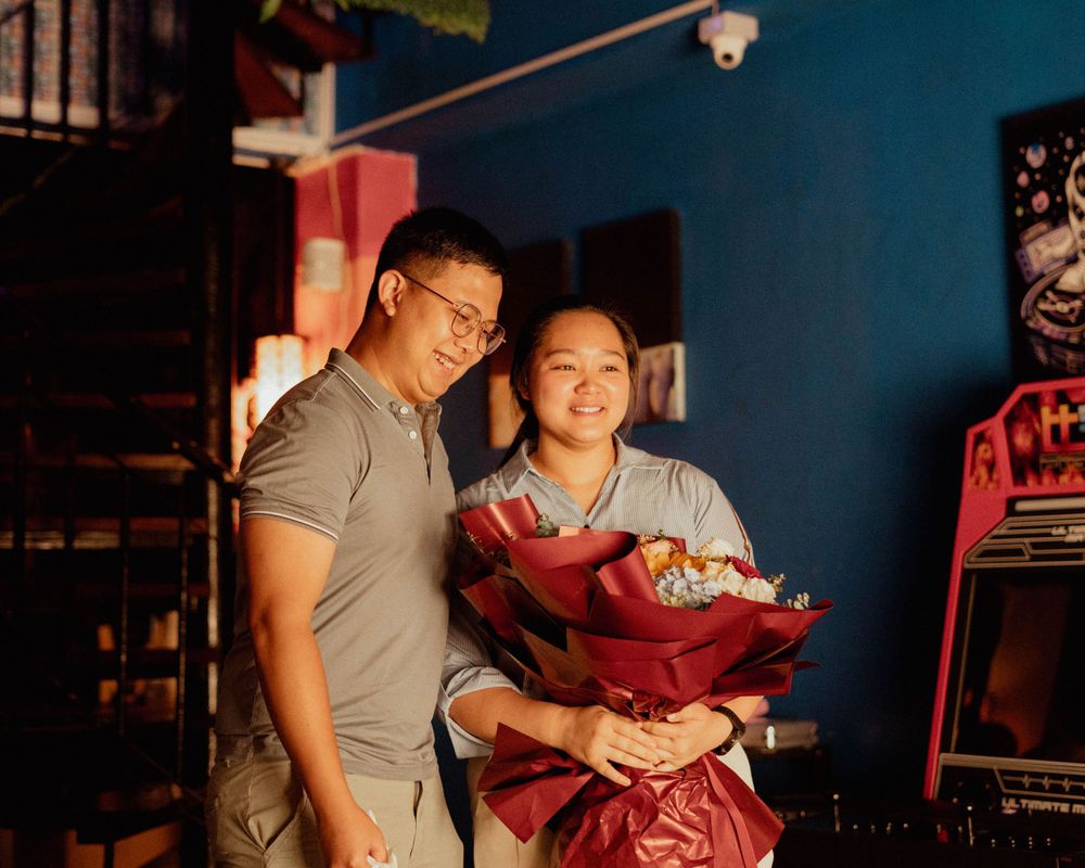 A couple smiles while holding a red bouquet in a warmly lit interior setting.