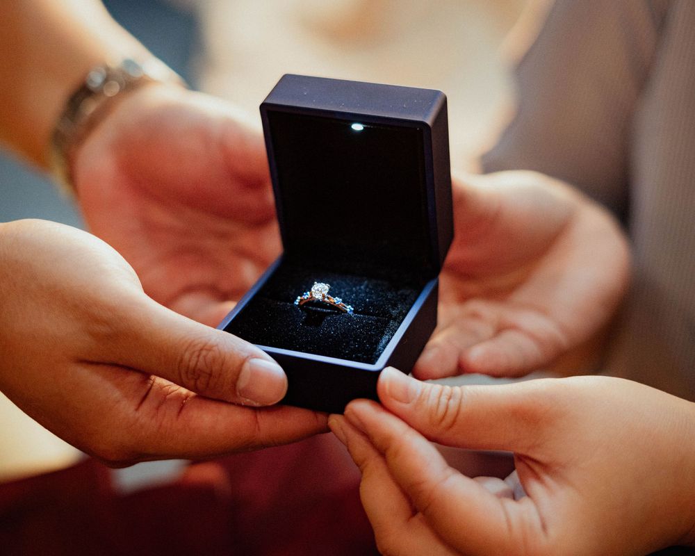 Hands presenting an engagement ring in a navy blue velvet box.