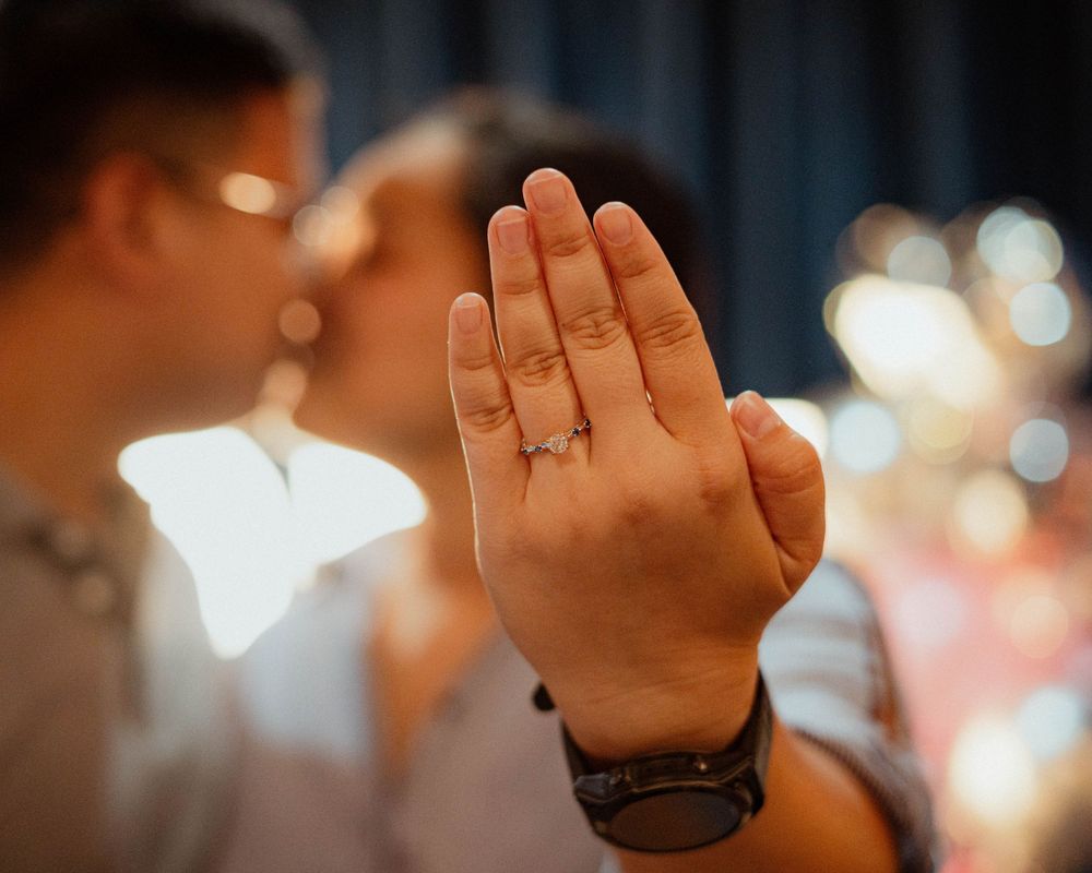 Hand displaying engagement ring against blurred romantic background.