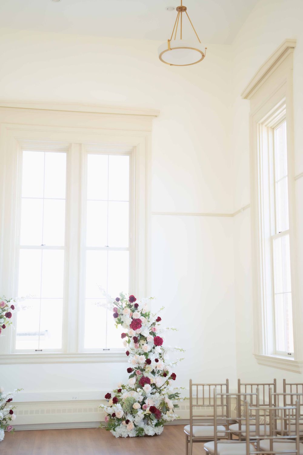 An elegant white room with tall windows featuring a floral arrangement of red and white flowers beside chiavari chairs.