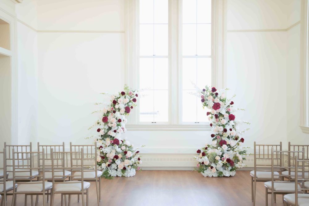 Wedding ceremony setup with symmetrical floral arrangements and gold chiavari chairs in a bright room. Location: Events at Ethereal.