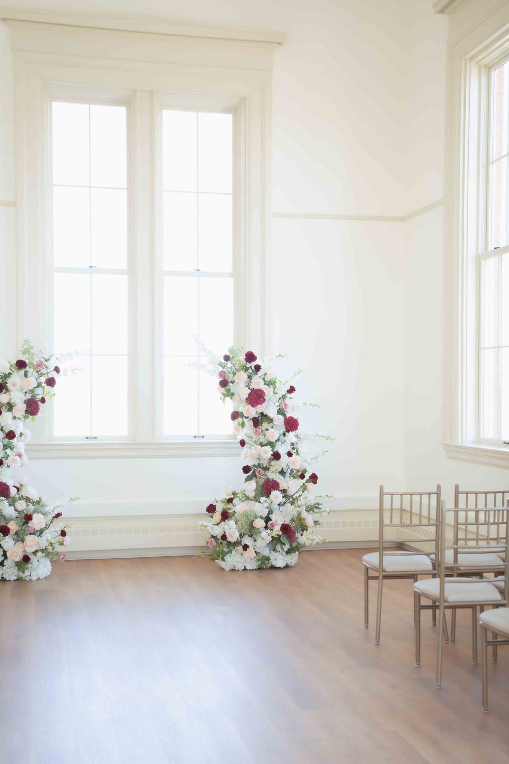 Minimalist wedding ceremony space with white walls and floral arrangements flanking windows.