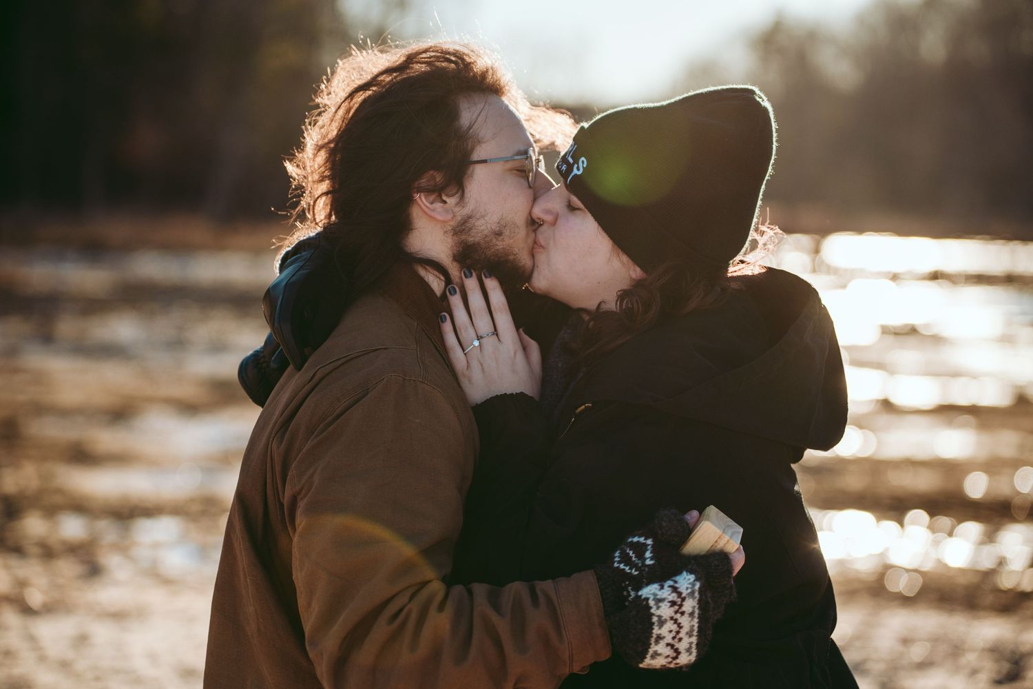 A couple shares an intimate kiss outdoors during golden hour sunlight with water and nature in the background.