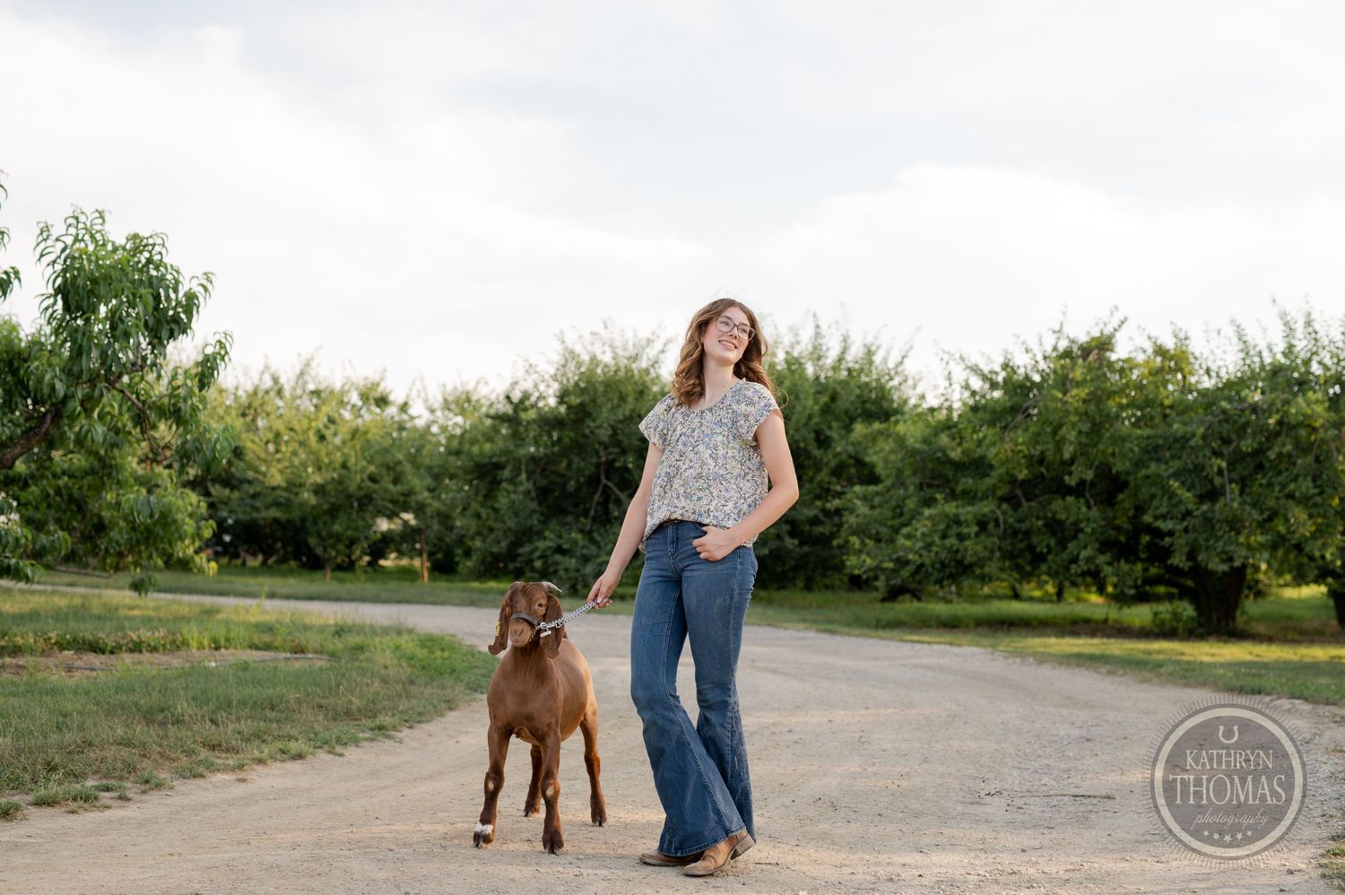 A brown goat and its handler walk together on a dirt path surrounded by greenery.